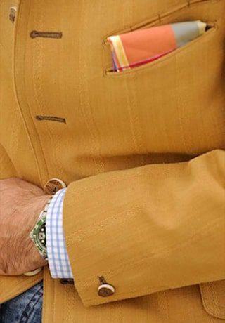 © Seisser & Cie. Muenchen GmbH Eine Person, die einen senfgelben Blazer mit braunen Knöpfen, ein gemustertes Einstecktuch und eine grüne Armbanduhr über einem blau-karierten Hemd trägt.