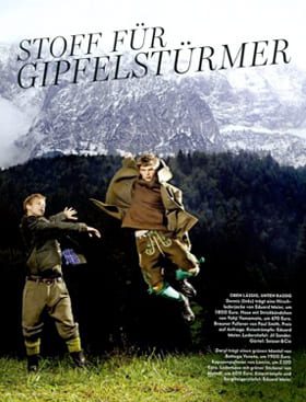 © Seisser & Cie. Muenchen GmbH Zwei Männer in traditioneller bayerischer Kleidung vollführen einen Sprung vor einer malerischen Kulisse aus schneebedeckten Bergen, Wald und bewölktem Himmel. Der deutsche Text lautet "Stoff für Gipfelstürmer" am oberen Rand.