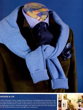 © Seisser & Cie. Muenchen GmbH Eine Schaufensterpuppe trägt eine braune Jacke, ein blaues Hemd, eine marineblaue gepunktete Krawatte und einen hellblauen Pullover, der über die Schultern gezogen ist. Der Kopf der Schaufensterpuppe ist mit einer bunten, gemusterten Kappe bedeckt.