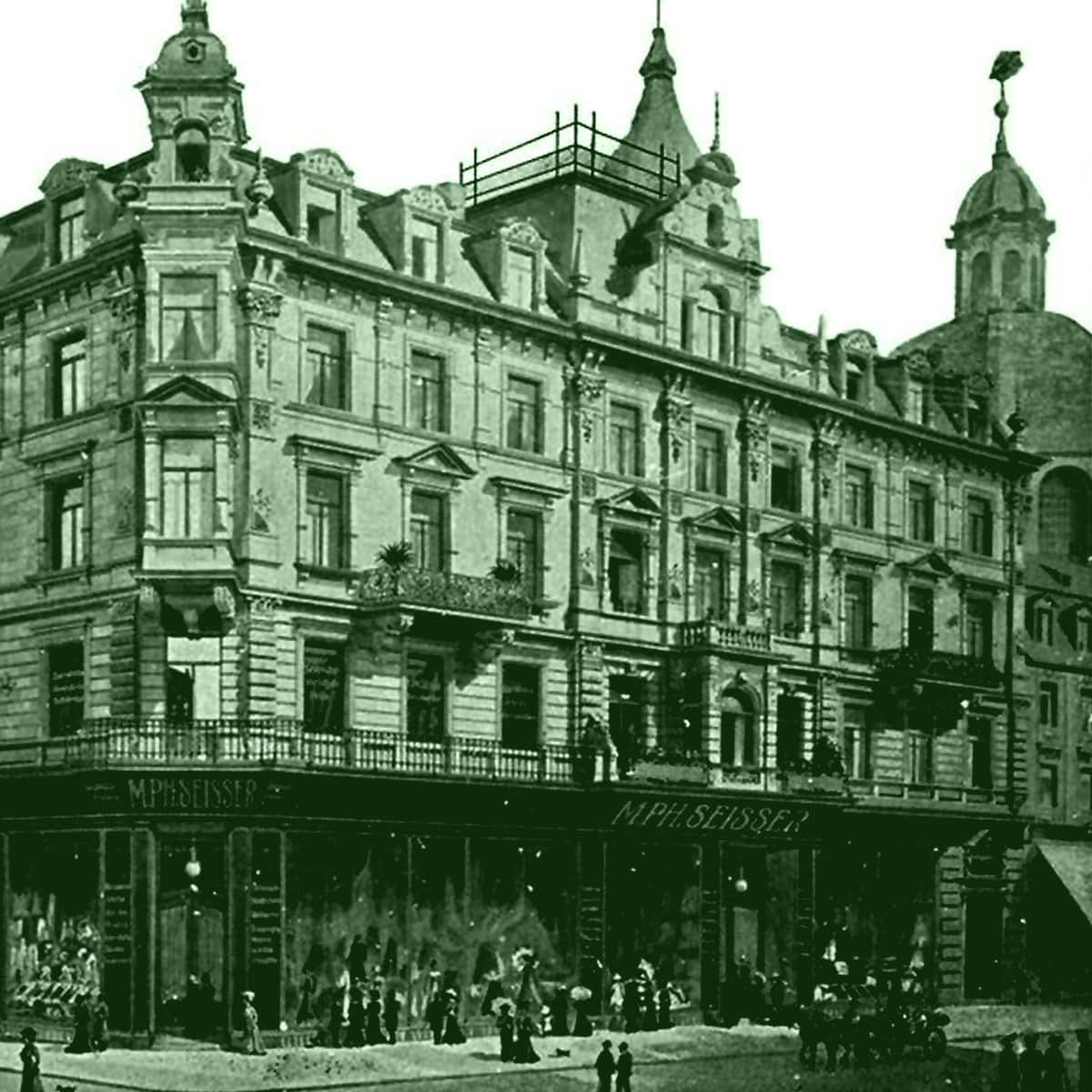 © Seisser & Cie. Muenchen GmbH Ein historisches mehrstöckiges Gebäude mit verschnörkelten architektonischen Details und Geschäften auf Straßenebene, darunter "M. P. Belser", mit Menschen, die sich draußen versammelt haben; im Hintergrund ist ein weiteres Hochhaus zu sehen. Das Bild hat einen Grünstich.
