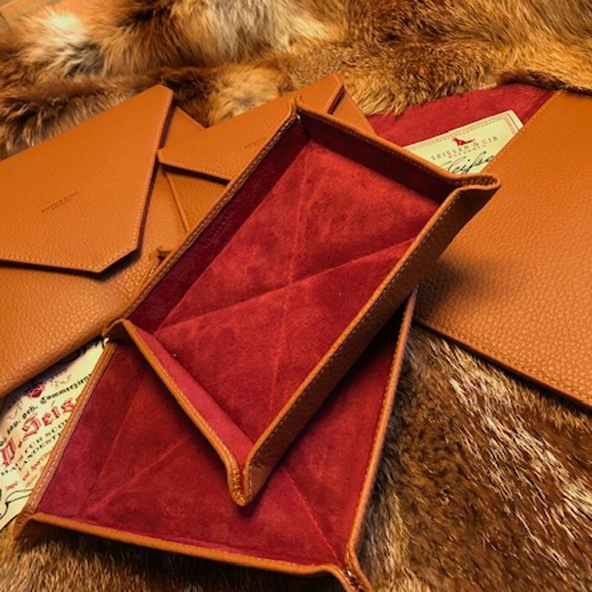 © Seisser & Cie. Muenchen GmbH Mehrere braune Ledertabletts und Briefumschläge mit roter Wildlederinnenseite sind auf einer Pelzoberfläche ausgestellt, wobei in einigen von ihnen Papiere im Vintage-Stil teilweise sichtbar sind.
