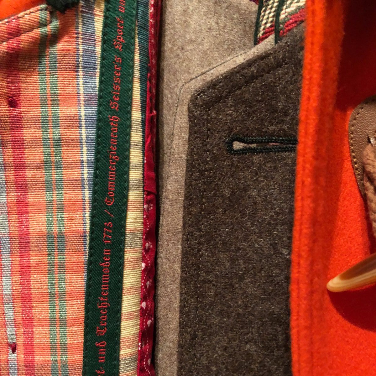 © Seisser & Cie. Muenchen GmbH Nahaufnahme der Innenseite eines Mantels, die einen bunt karierten Stoff, graue Wolle und orangefarbenes Material zeigt. Am Rand des karierten Futters ist ein grüner Text in deutscher Sprache aufgestickt. Ein Holzbügel ist teilweise sichtbar.