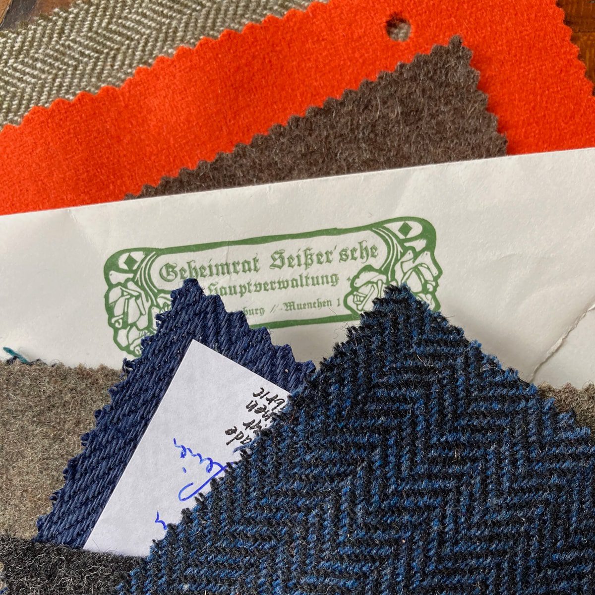 © Seisser & Cie. Muenchen GmbH Mehrere Stoffmuster in verschiedenen Farben und Mustern, darunter Grau, Blau, Orange und Fischgrät, sind übereinandergelegt. Unter den Stoffen befindet sich ein teilweise sichtbarer Umschlag mit verschnörkeltem deutschen Text.