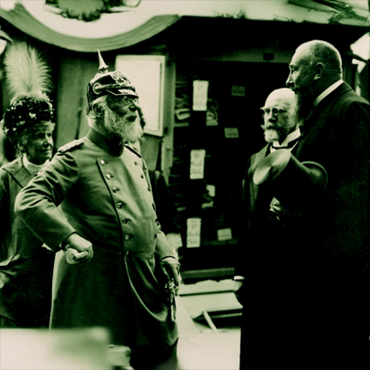 © Seisser & Cie. Muenchen GmbH Vier Männer in historischen Gewändern stehen zusammen und unterhalten sich. Einer trägt eine verzierte Militäruniform mit Stachelhelm, die anderen formelle Anzüge; einer hat einen Federhut. Der Schauplatz scheint im Freien zu sein, möglicherweise bei einer Veranstaltung oder Versammlung.
