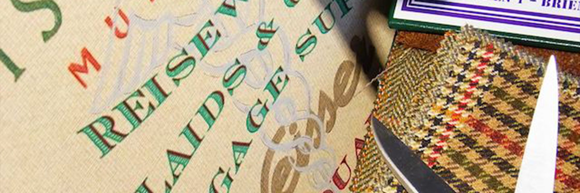 © Seisser & Cie. Muenchen GmbH Nahaufnahme eines Stoffes mit den Wörtern "REISE", "LUGGAGE" und "SUIT" in Grün und Rot, daneben eine Schere, gemusterter Tweed-Stoff und ein Etikett, das in der oberen rechten Ecke teilweise sichtbar ist.