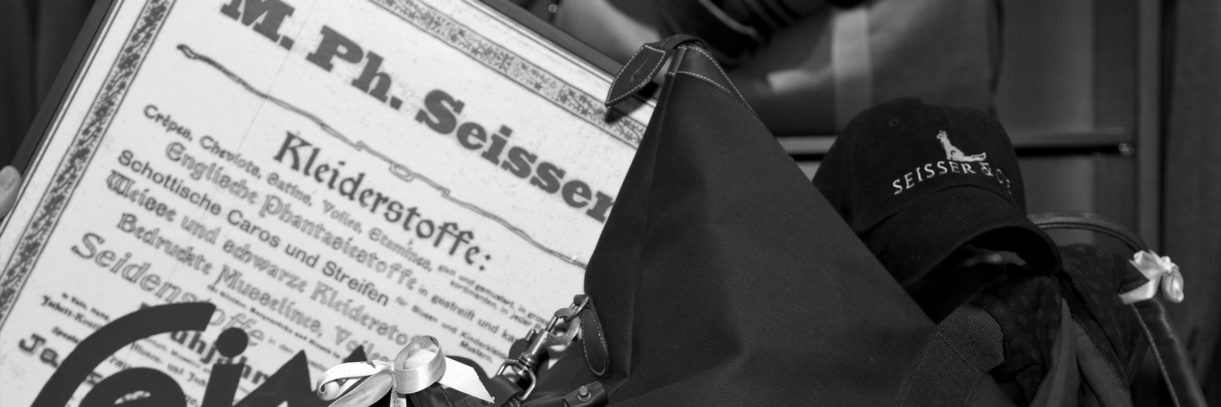 © Seisser & Cie. Muenchen GmbH Ein Schwarz-Weiß-Foto, das eine gerahmte Vintage-Werbung in deutscher Sprache, eine Handtasche mit Schleifenverzierung und eine Mütze mit dem teilweise sichtbaren Logo "SEISSER" zeigt.