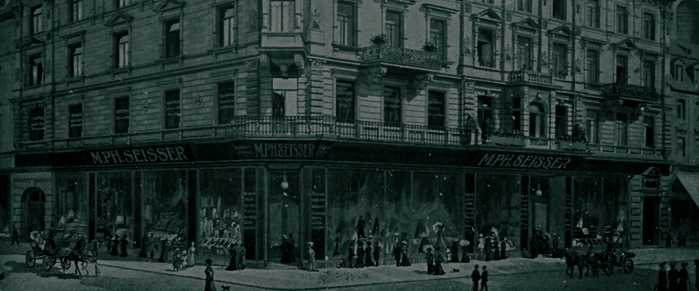 Historisches Foto eines großen Eckgebäudes mit verschnörkelter Architektur; im Erdgeschoss befinden sich "MPH. SEISSER"-Schilder und große Schaufenster. Menschen und Pferdekutschen sind draußen versammelt.