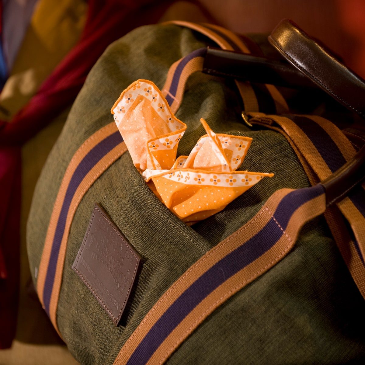 © Seisser & Cie. Muenchen GmbH Nahaufnahme einer grün und braun gestreiften Reisetasche mit zwei orange gemusterten Origami-Booten in der Vordertasche. Das Licht schafft eine warme, gemütliche Atmosphäre.