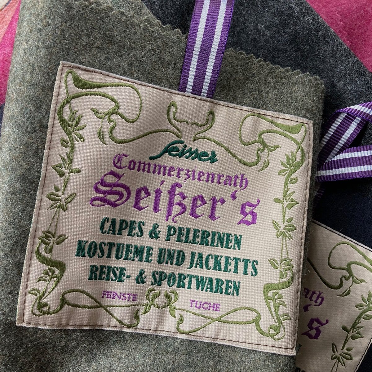 © Seisser & Cie. Muenchen GmbH Ein Stoffetikett mit violettem und grünem Text in deutscher Sprache, das für Umhänge, Jacken, Kostüme und Reisekleidung von "Seiker" wirbt, umrandet von einem verschnörkelten grünen Muster und mit einem violett gestreiften Band an einem Wollstoff befestigt.