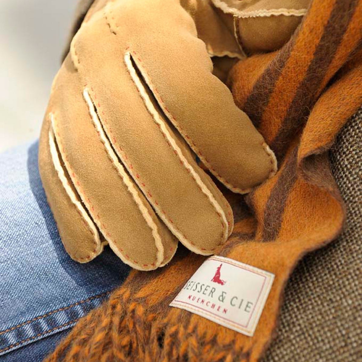 © Seisser & Cie. Muenchen GmbH Nahaufnahme eines hellbraunen Wildlederhandschuhs, der auf einem Wollschal mit braunen und orangefarbenen Streifen ruht und ein Etikett mit der Aufschrift "HEISSER & CIE MÜNCHEN" zeigt. Ein Teil einer Tweedjacke und eine blaue Jeans sind ebenfalls zu sehen.