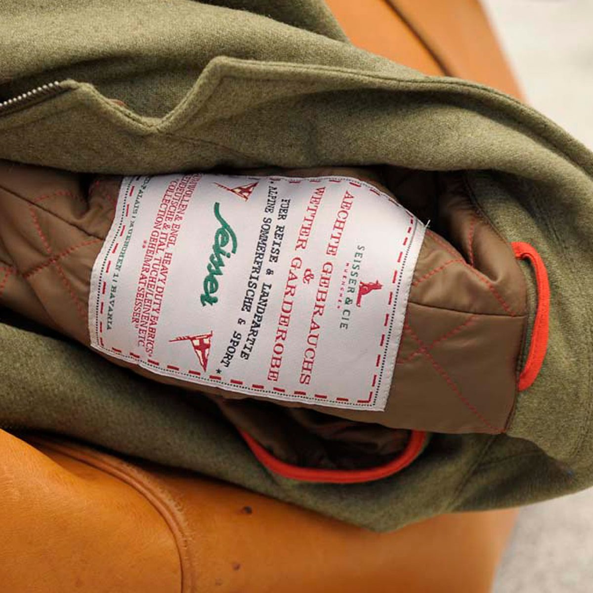 Nahaufnahme der Innenseite einer olivgrünen Jacke mit einem großen weißen Etikett mit rotem Text und Pflegehinweisen in deutscher Sprache, das teilweise auf ein gestepptes braunes Futter mit roten Nähten genäht ist.